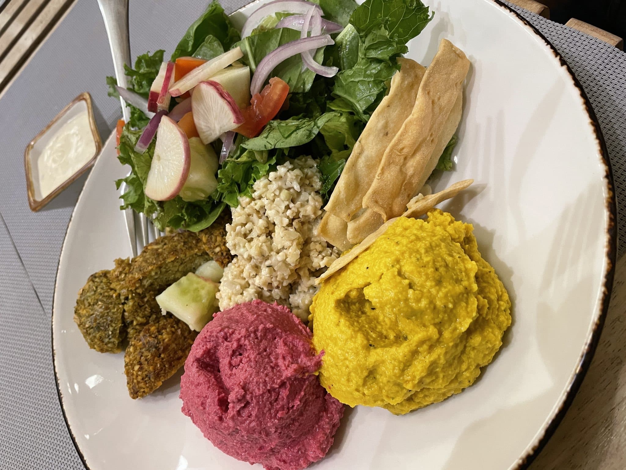 Plato de ensalada verde con bulgur, falafel, hummus de remolacha y una salsa amarilla.