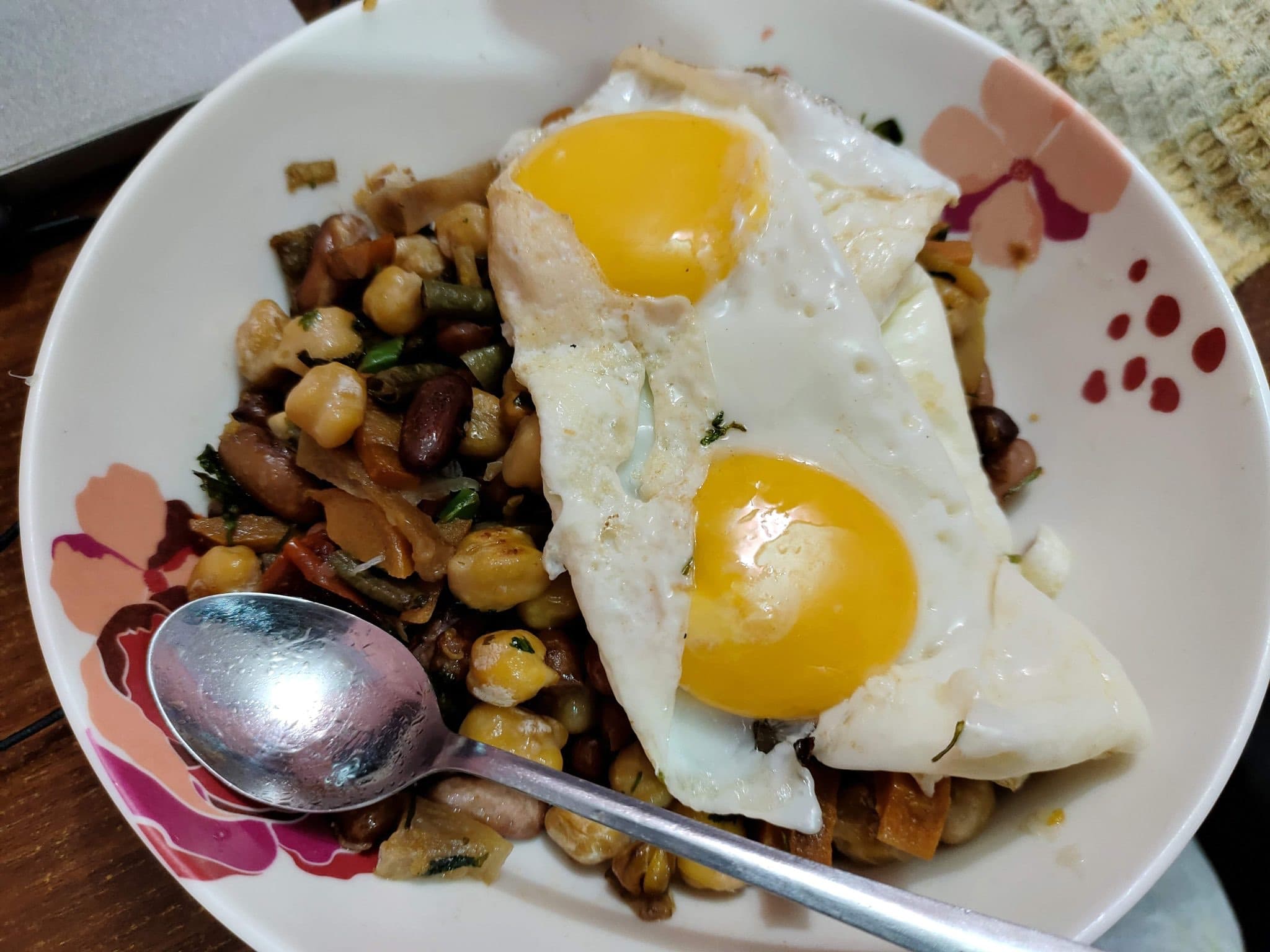 Un bol de alubias y garbanzos con dos huevos fritos encima y una cuchara al lado.