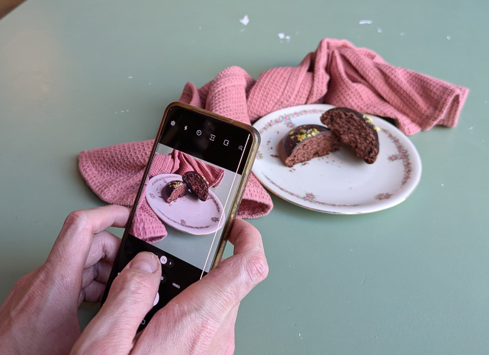 Dos manos sostienen un móvil y fotografían dos mitades de una tarta de chocolate sobre un plato de flores.